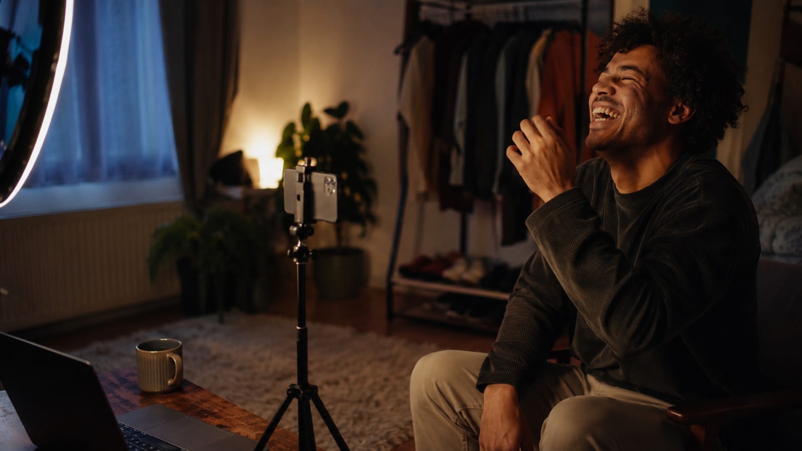 A creator sitting in a warmly lit home living room laughing at a flubbed take, with a phone on a tripod filming him and a ring light at the edge of the frame