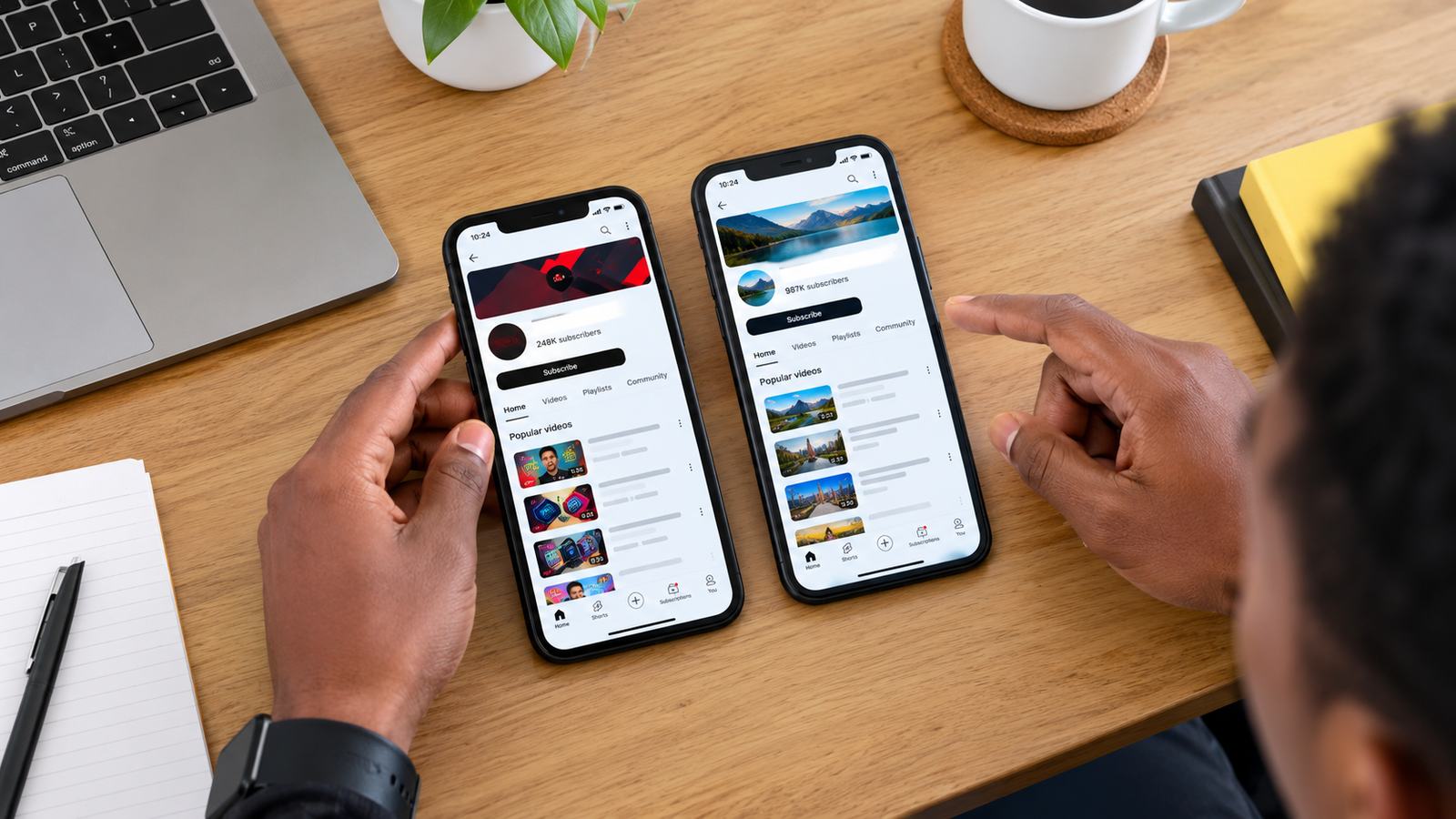 Overhead view of two phones side by side on a desk, each showing a different YouTube channel page with subscriber counts, hands comparing them